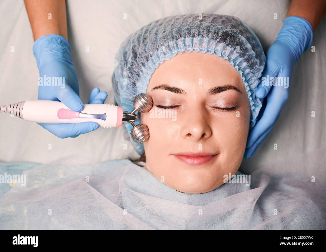 Gros plan des mains des cosmétologues à l'aide d'un rouleau facial anti-âge à microcourant tout en massant le visage des femmes clientes. Jeune femme en bonnet de douche jetable ayant un massage galvanique dans le salon de beauté. Banque D'Images