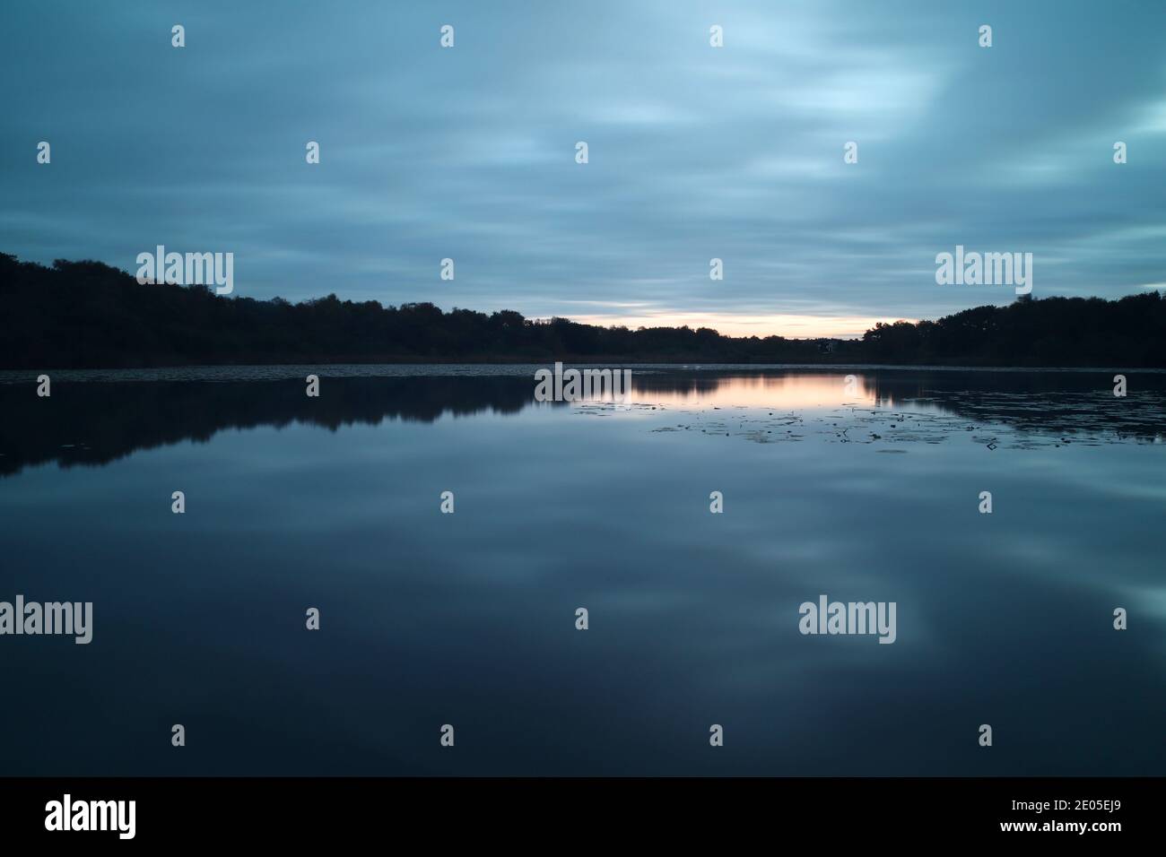 Une prise de vue à longue exposition prise avant le lever du soleil capture une scène de mauvaise humeur aux tons subdués de bleu et de gris reflétés dans l'eau fixe de Hatch Pond Banque D'Images