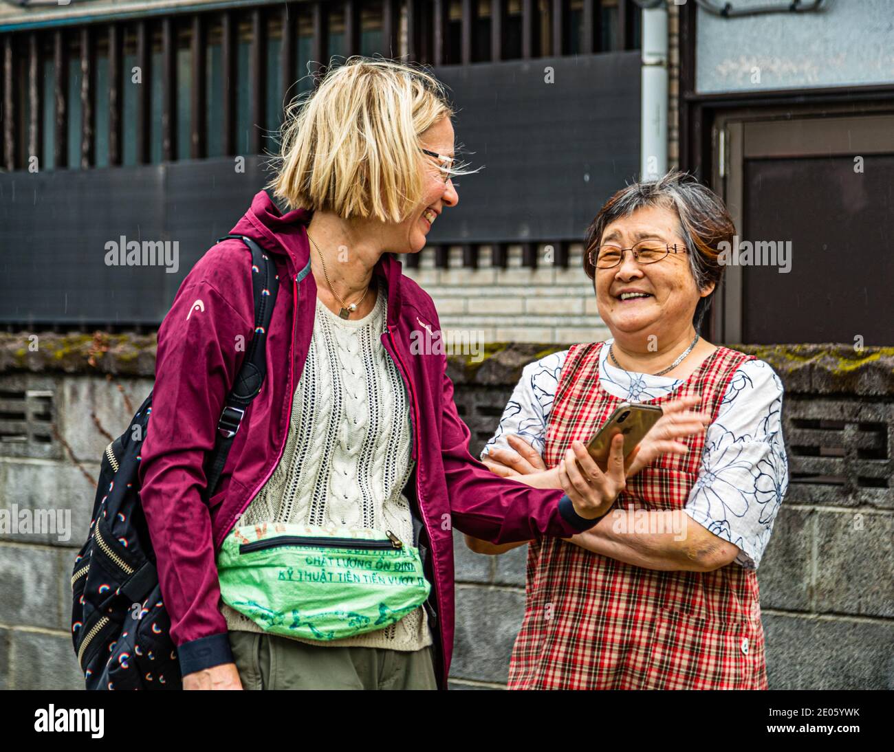 La journaliste de voyage Angela Berg et Mme Iyima communiquent à l’aide d’une application de traduction sur leurs smartphones devant un bâtiment en bois traditionnel à Nishiizu-Cho, préfecture de Shizuoka, au Japon – un ‚choc culturel‘ que les deux résolvent avec un rire, grâce à la traduction, alors qu’une légère pluie tombe. Banque D'Images