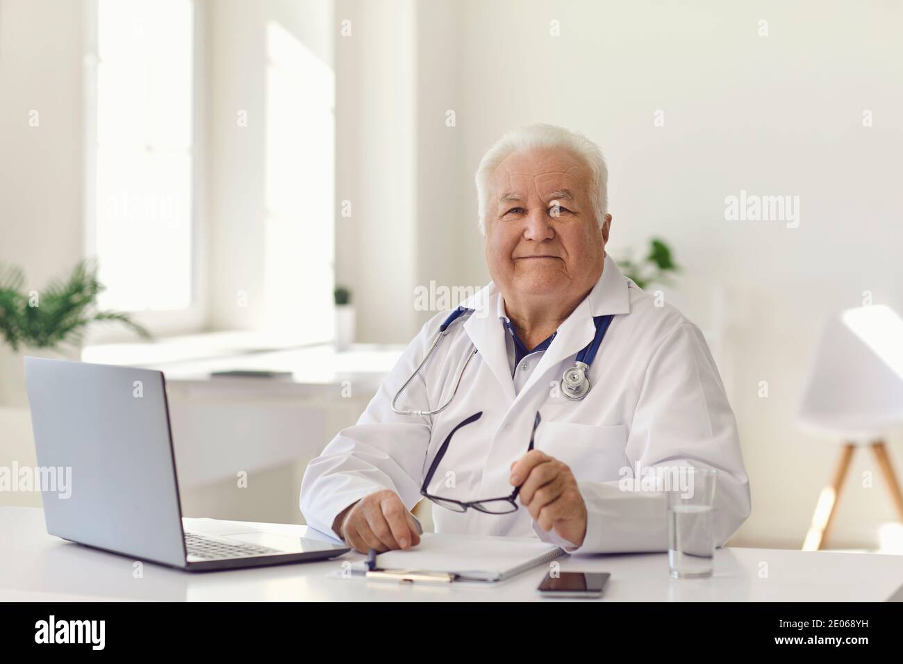 Médecin expérimenté sympathique assis à un bureau avec un ordinateur portable dans un cadre moderne bureau de l'hôpital et regarder la caméra Banque D'Images