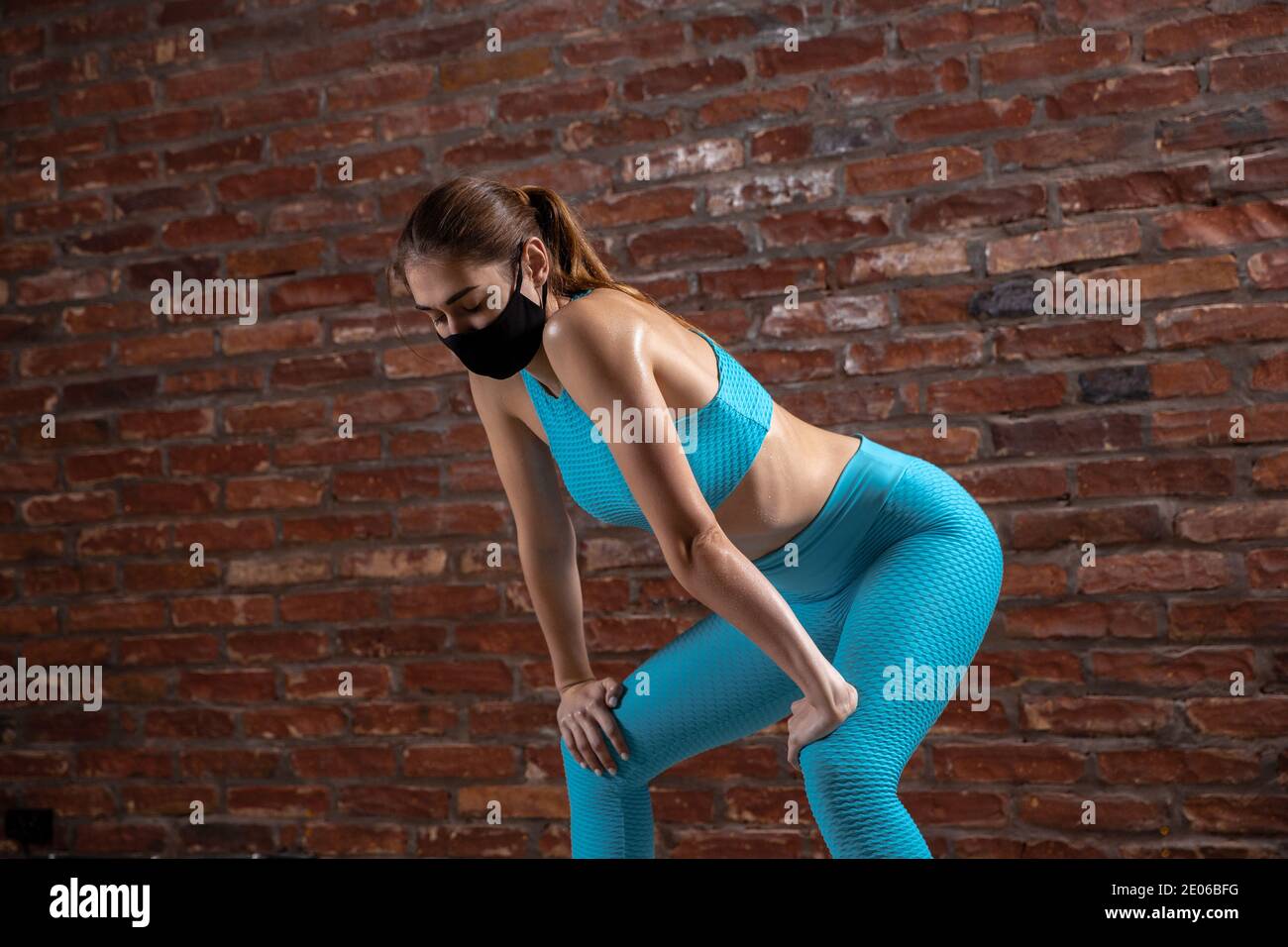 Ajuster. Entraînement professionnel de l'athlète féminine sur fond de mur de brique portant un masque facial. Sport pendant la quarantaine du coronavirus dans le monde. Jeune femme pratiquant dans la salle de gym en toute sécurité en utilisant l'équipement. Banque D'Images