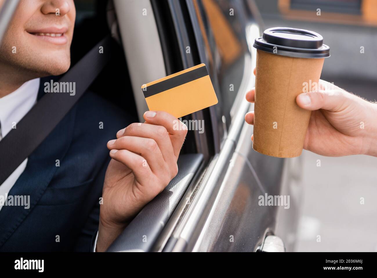 Vue rognée d'un homme d'affaires souriant dans une voiture tenant une carte de crédit proche vendeur avec café à emporter Banque D'Images