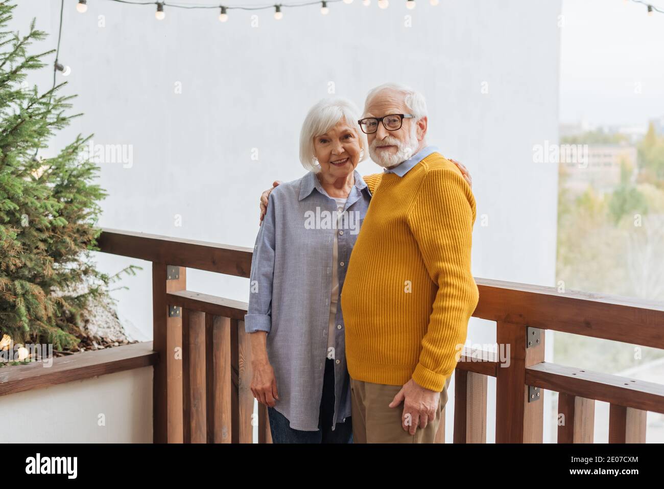 couple senior souriant regardant l'appareil photo tout en s'embrassant sur la terrasse près du pin sur fond flou Banque D'Images