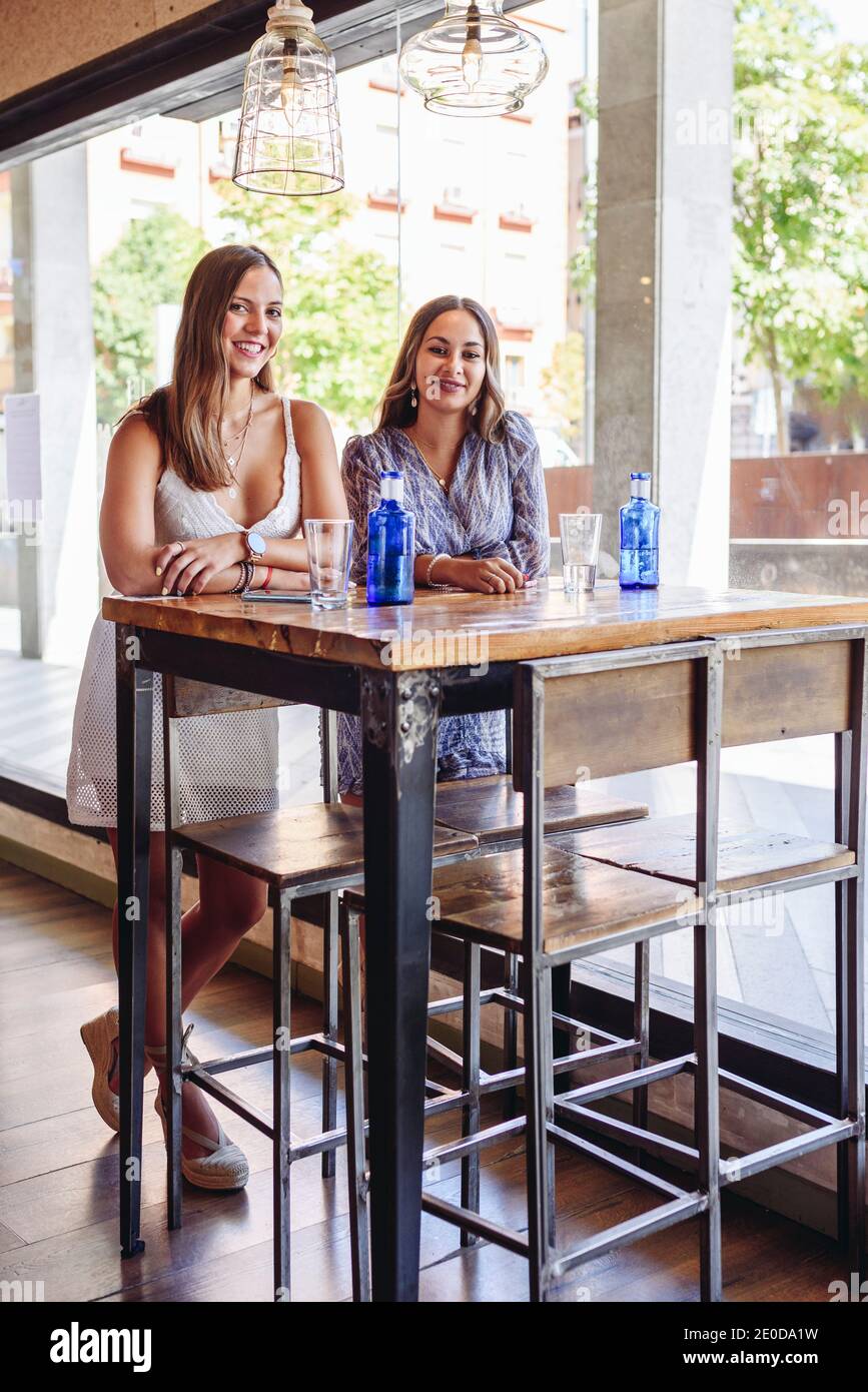 Jeunes amies souriantes regardant l'appareil photo tout en se tenant debout à la table avec boissons pendant la réunion dans la cafétéria moderne en été Banque D'Images