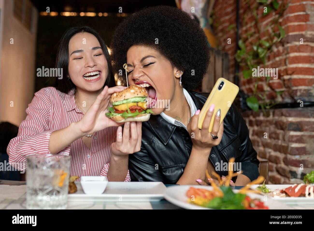 Drôle de jeune Afro-américaine femme prenant le selfie sur smartphone tout en piquant délicieux hamburger tenu par rire asiatique petite amie pendant le dîner togeth Banque D'Images