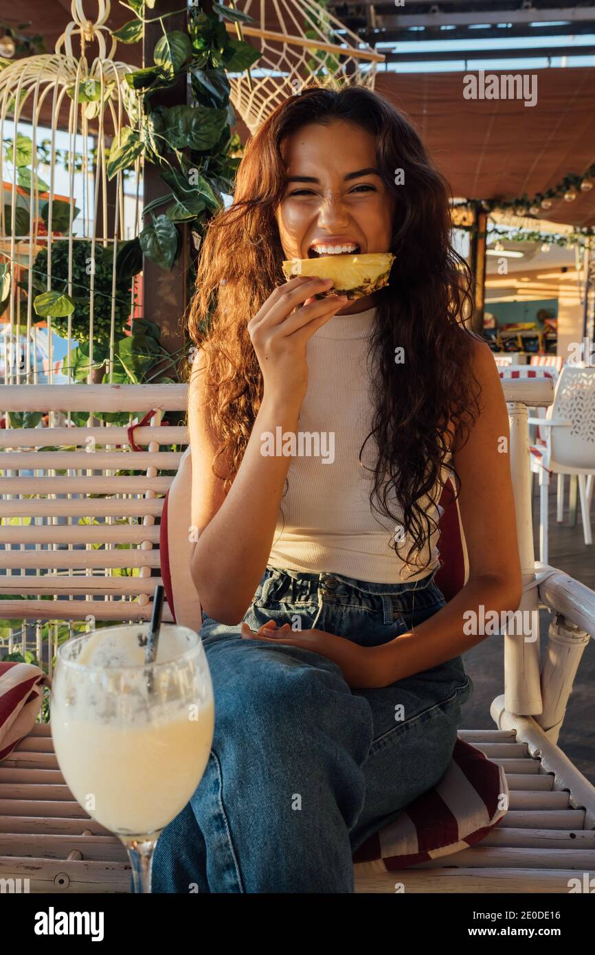 Une jeune femme ludique fait de la grimace et mord de l'ananas frais pendant se rafraîchir dans un café en plein air en soirée d'été Banque D'Images