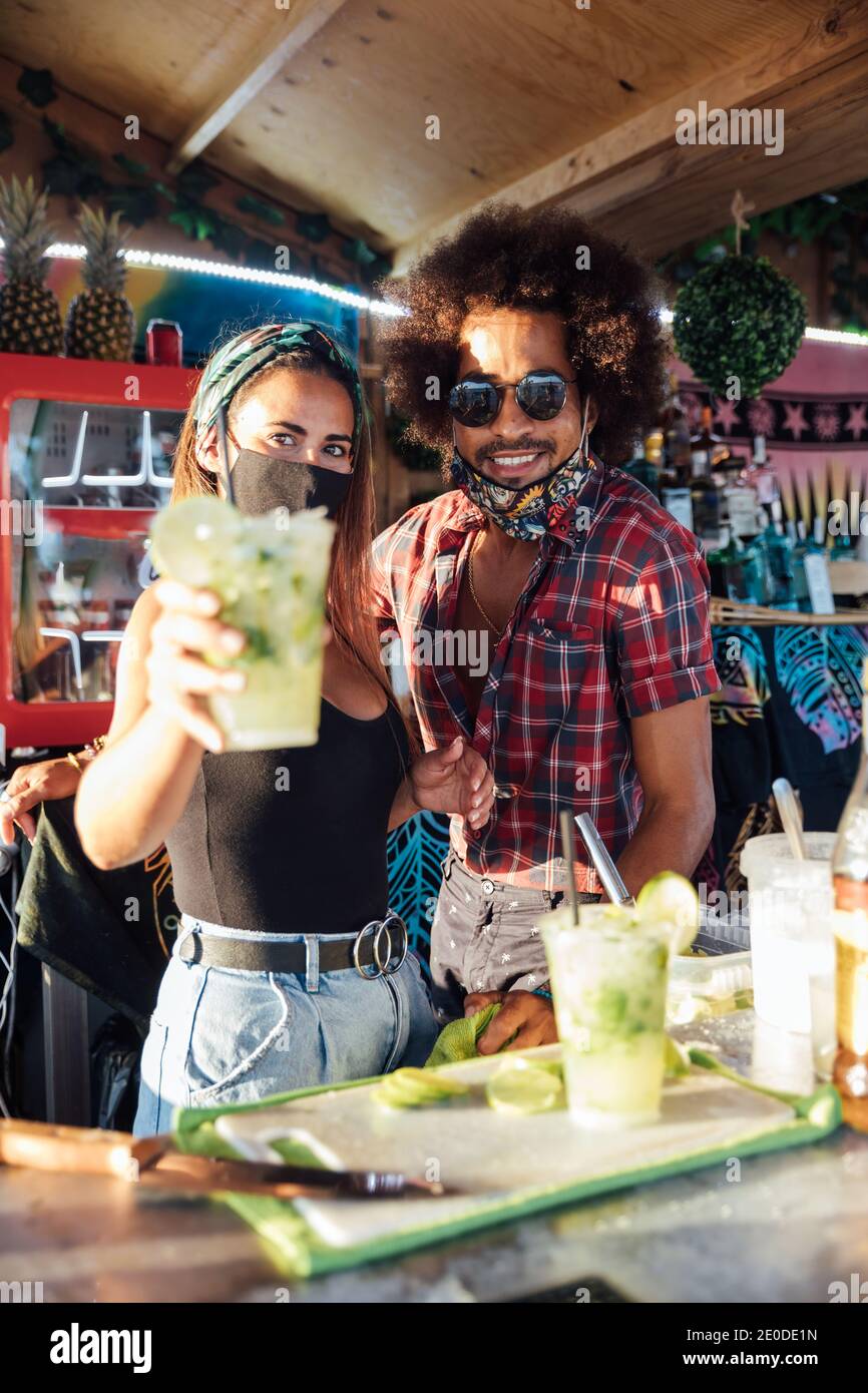 Les propriétaires ethniques positifs du bar tropical regardant l'appareil-photo amical et propose un verre de cocktail rafraîchissant avec du citron vert et de la glace Banque D'Images