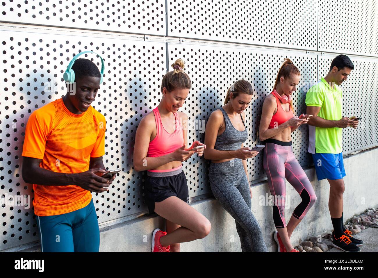 Groupe de coureurs multiraciaux en vêtements d'activité penchée sur le mur de construire et utiliser des smartphones après un entraînement en ville Banque D'Images