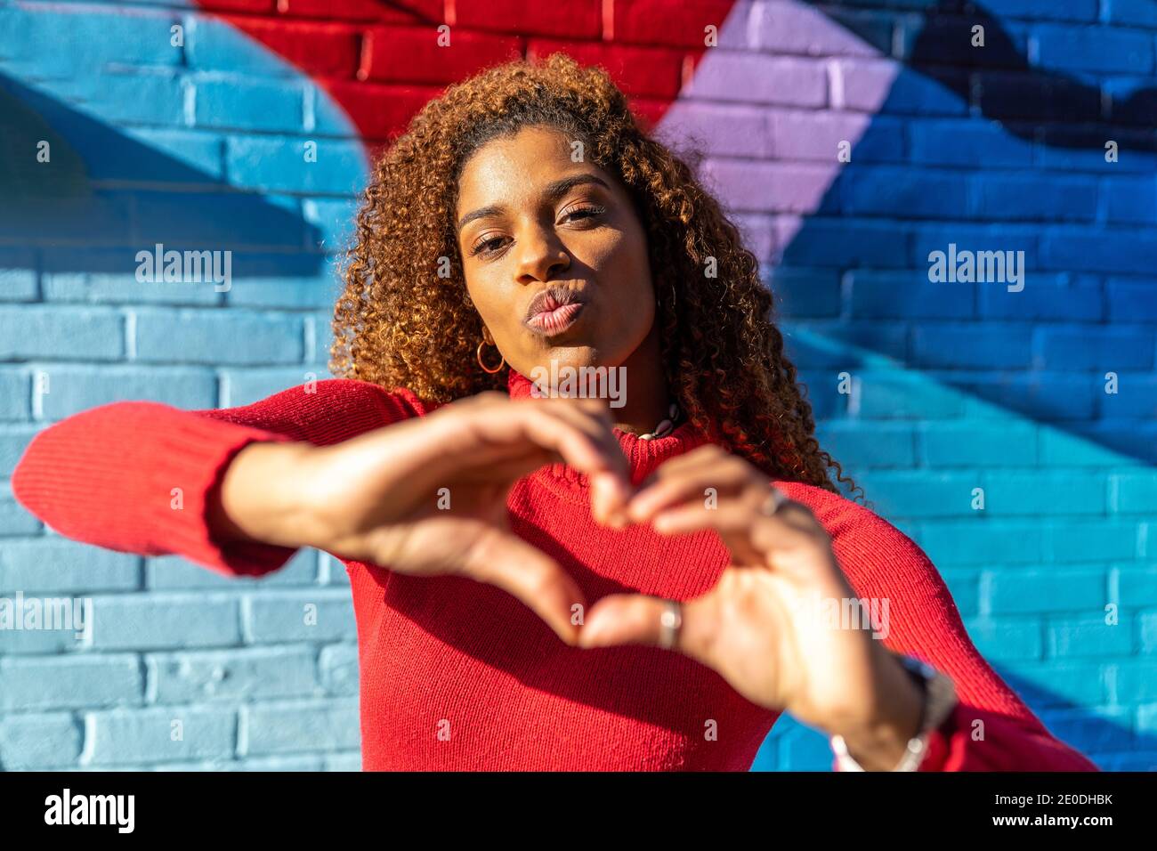 Jeune femme ethnique confiante avec cheveux afro dans un vetement decontracte faire des pieds sur les levres et montrer le signe du coeur avec les mains tout en etant debout dans la
