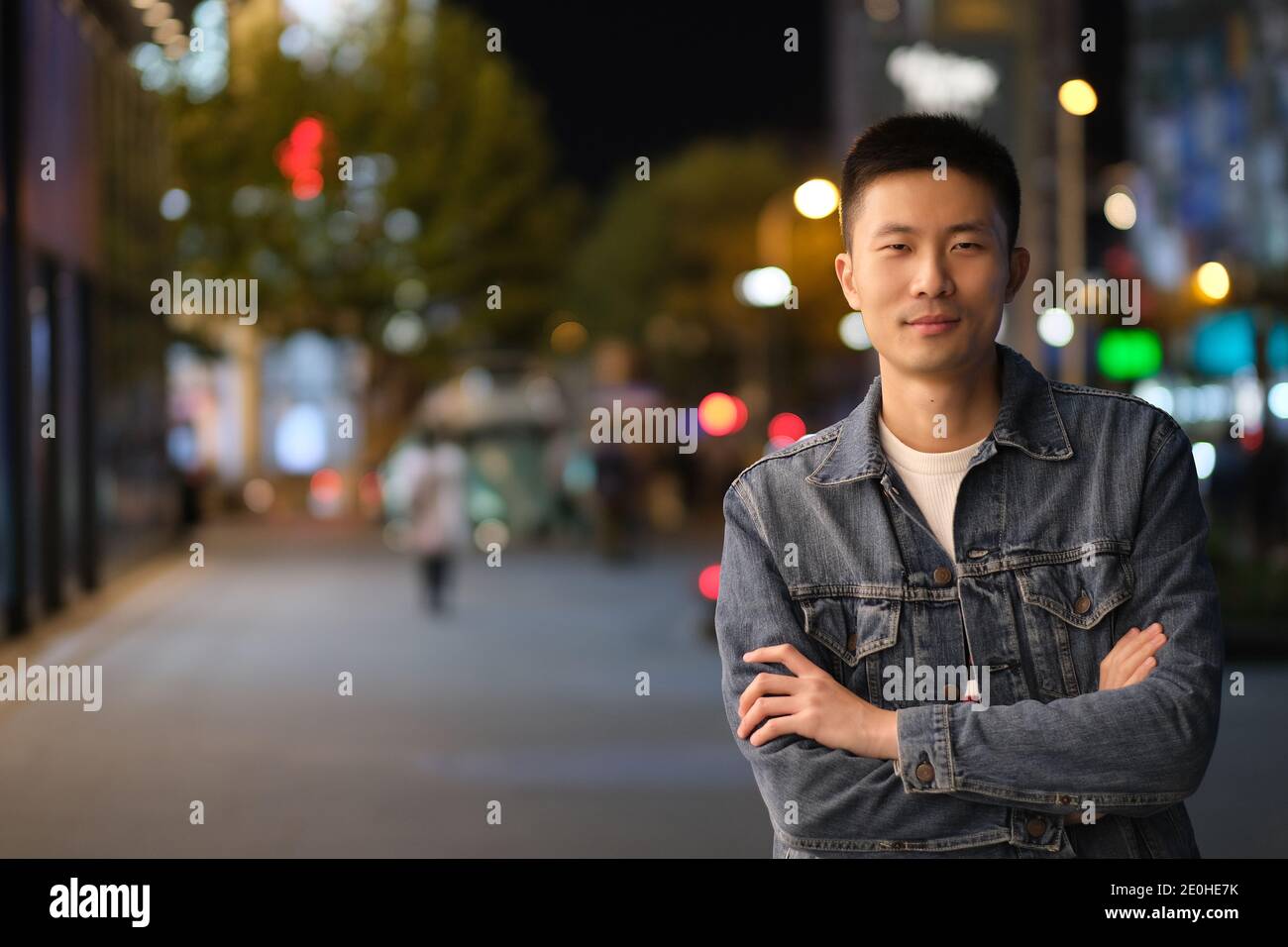 Photo d'un jeune asiatique de nuit dans une ville moderne, bras croisés et souriant, en regardant l'appareil photo Banque D'Images