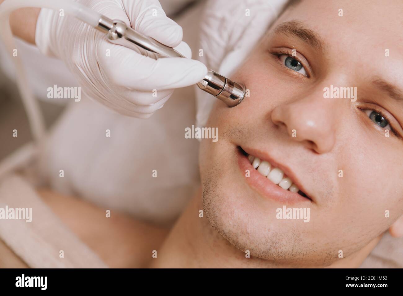 Petit gros plan d'un jeune homme joyeux souriant pendant un cosmétologue qui effectue un traitement de soin du visage Banque D'Images