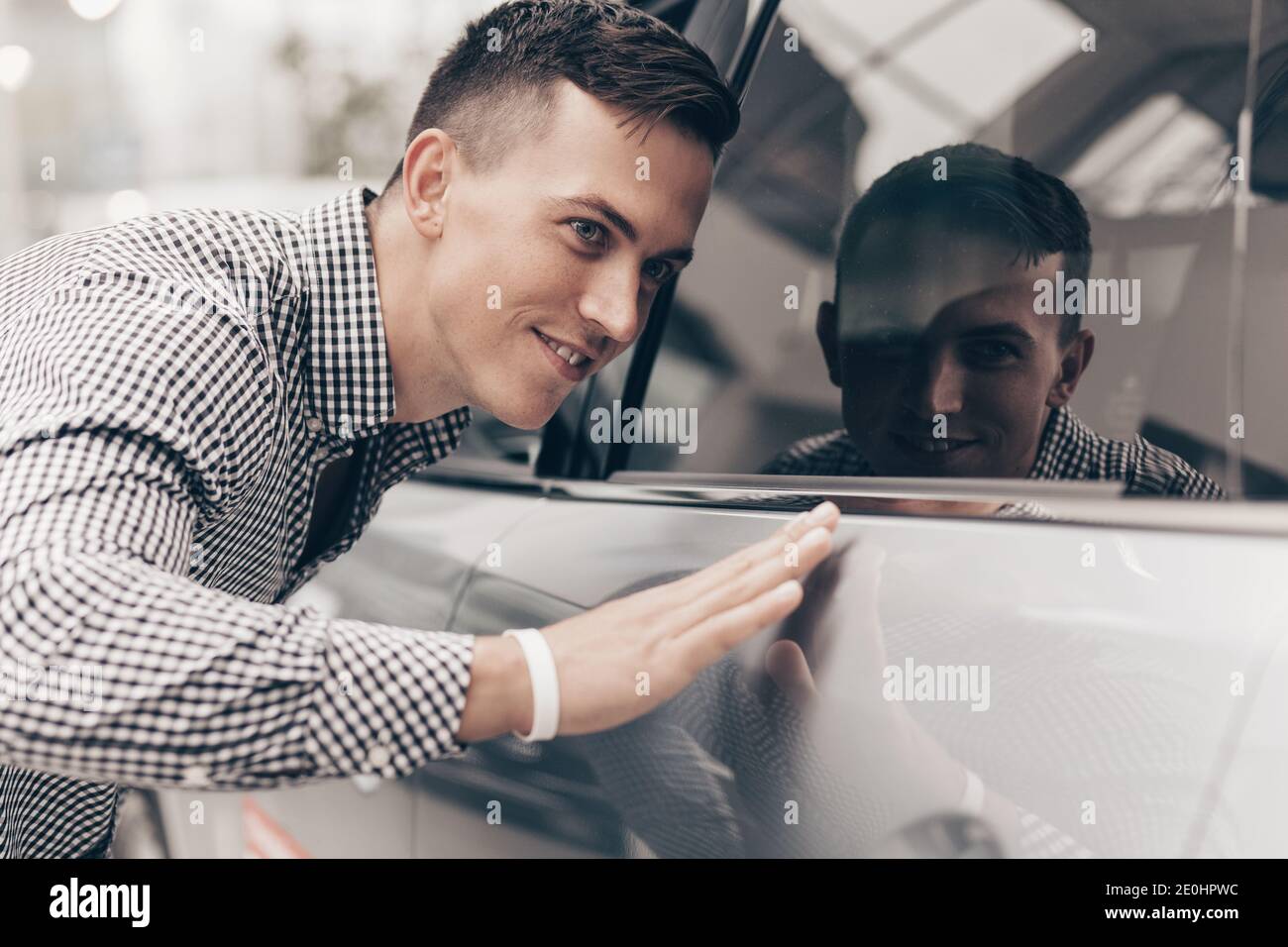 Gros plan d'un bel homme heureux touchant doucement la nouvelle voiture à la concession. Un client masculin attractif examine les voitures en vente au salon automobile showroo Banque D'Images