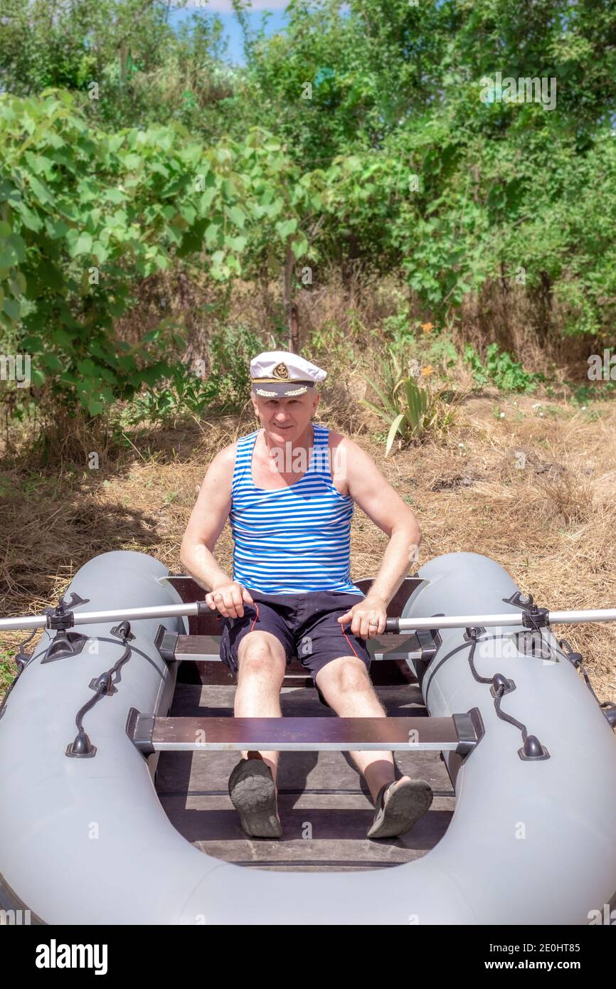 un homme dans une casquette navale est assis dans un bateau en caoutchouc  debout sur l'herbe. Un marin à la retraite manque la mer. Entretien et  fonctionnement du bain gonflable en caoutchouc