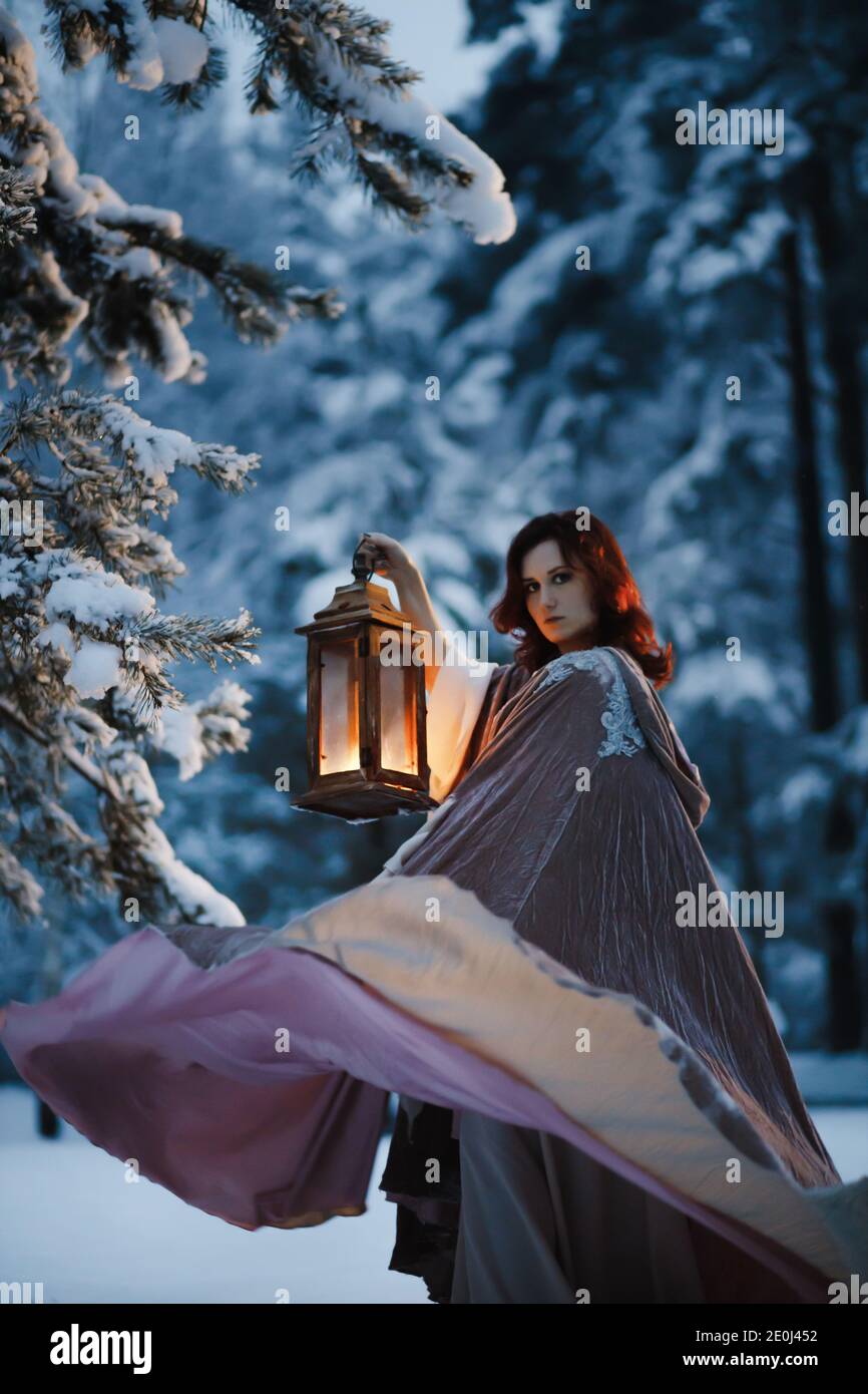 Photo de fantaisie épique : jeune femme qui tourne son manteau volant avec une lanterne vintage dans une forêt enneigée la nuit. Concept cosplay médiéval de conte de fées Banque D'Images