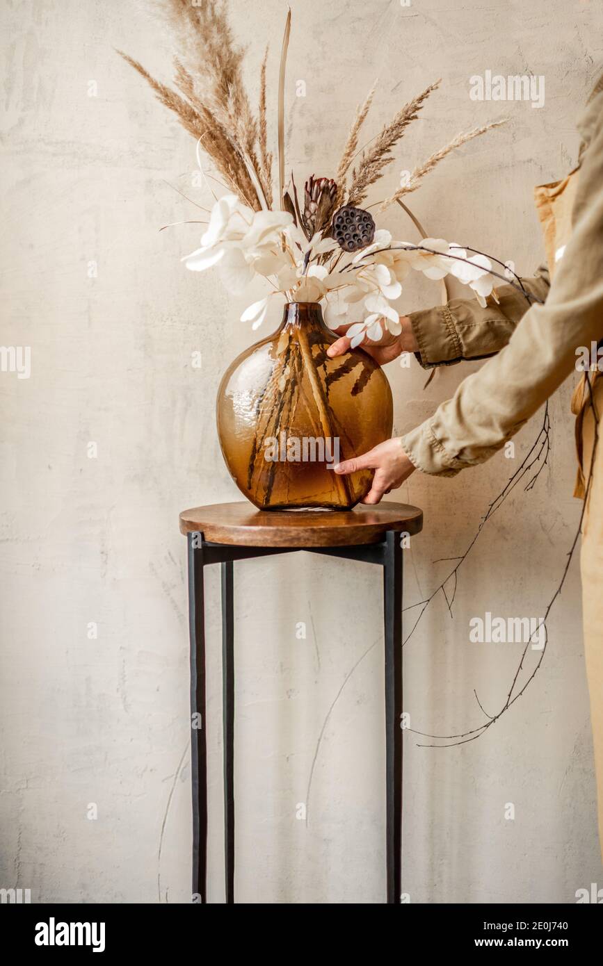Femme décorant la maison avec une composition de fleurs séchées et herbes dans un vase en verre sur un fond de mur beige Banque D'Images