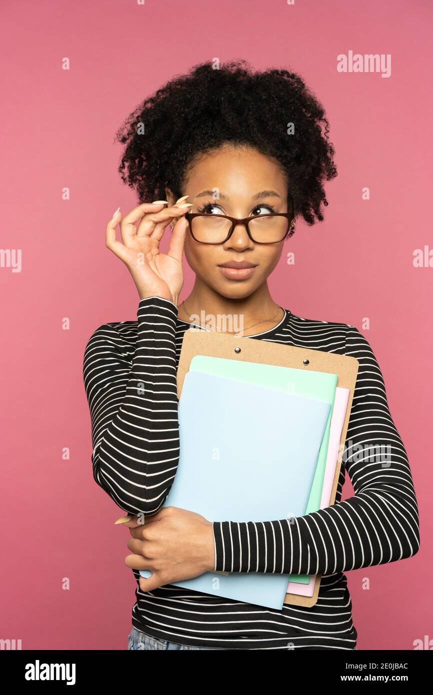 Femme enseignante ou tutrice afro-américaine isolée sur un mur rose de studio. Fille étudiante touchant des lunettes, tenant des carnets, regardant de côté. Éducation Banque D'Images