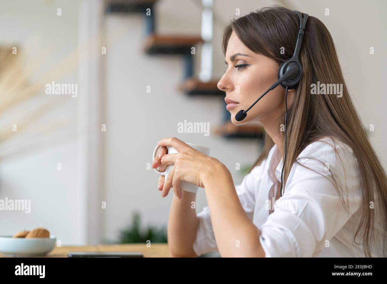 Une femme attentionnés, assise sur une table, regardant un webinaire, portant un casque, communiquant avec le client via un appel vidéo sur ordinateur, prenant du thé Banque D'Images