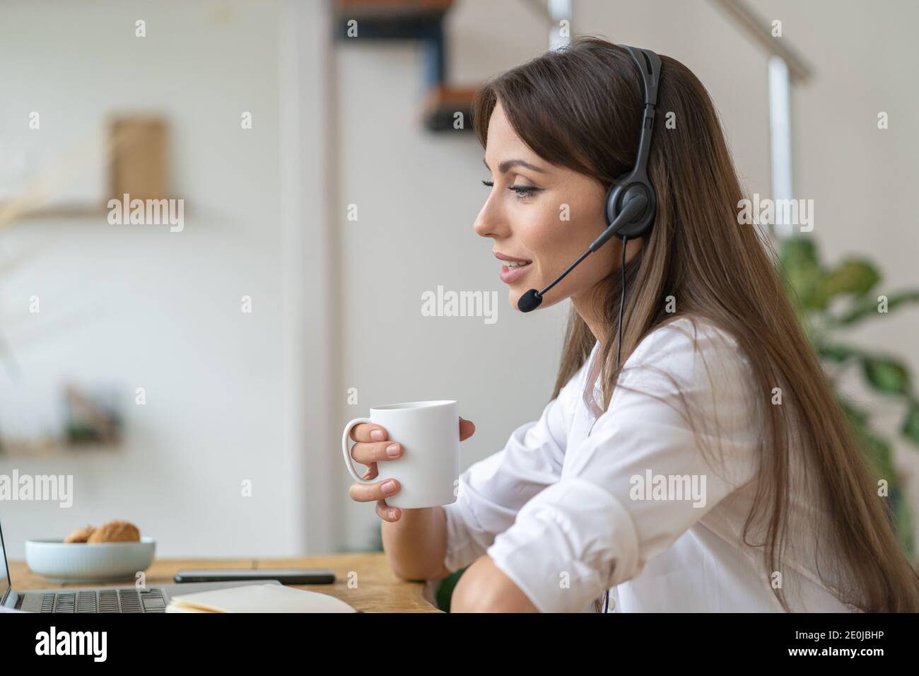 Femme d'affaires assise sur une table, participe à la vidéoconférence, porte des écouteurs, communique avec le client par vidéoconférence, boire du thé. Banque D'Images