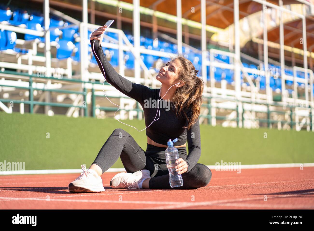 Installez une femme dans un vêtement de sport, reposez-vous après l'entraînement ou la course, asseyez-vous sur le circuit du stade, prend un selfie, écoute de la musique avec des écouteurs filaires sur smartpho Banque D'Images