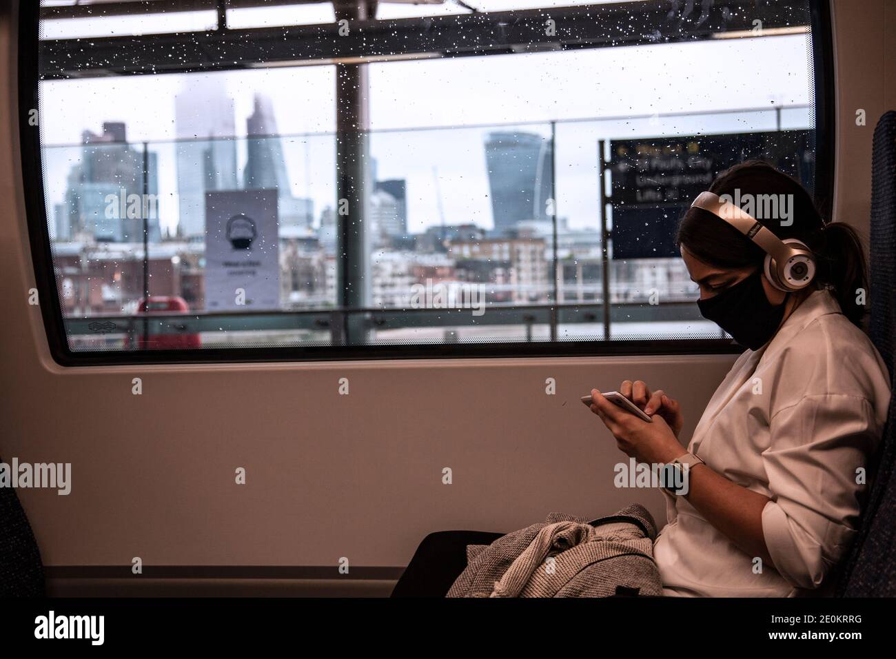 Une jeune femme dans un train portant un masque dactylographiant sur son téléphone avec la vue du quartier financier en arrière-plan. Londres Banque D'Images