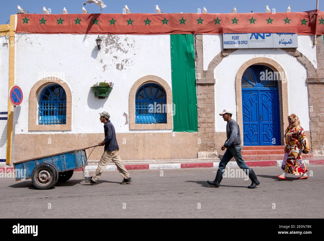 Les travailleurs marchent le long d'une rue au port de pêche d'Essaouira, sur la ville de l'océan Atlantique au Maroc, tandis que des mouettes bordent le toit d'un bâtiment. Banque D'Images