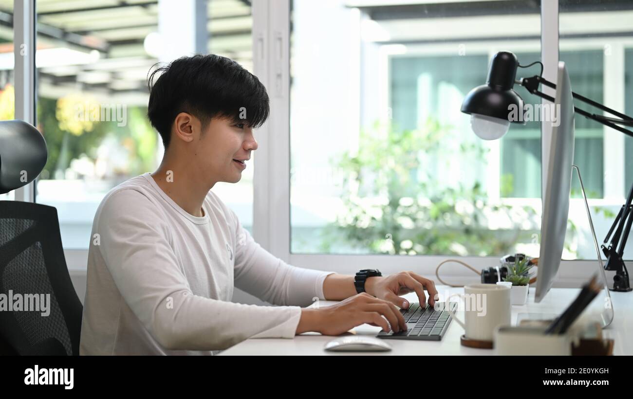 Vue latérale de l'homme indépendant en vêtements décontractés assis dans le bureau à la maison et travaillant sur le projet avec ordinateur. Banque D'Images
