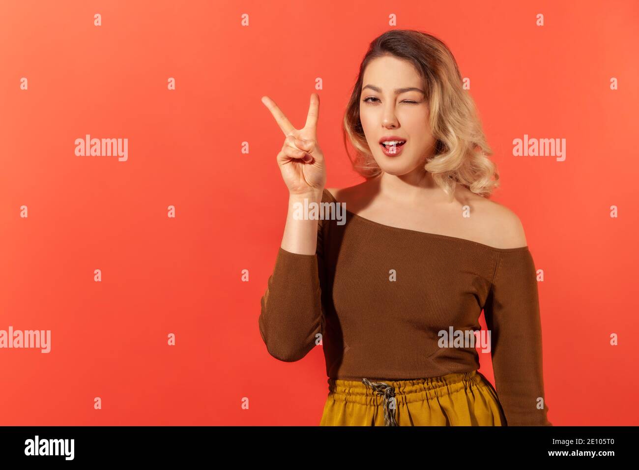 Portrait d'une jeune femme heureuse avec des cheveux blonds dans un chemisier marron montrant le geste de victoire par ses doigts et en se faisant un clin d'œil à l'appareil photo. Goujon intérieur Banque D'Images
