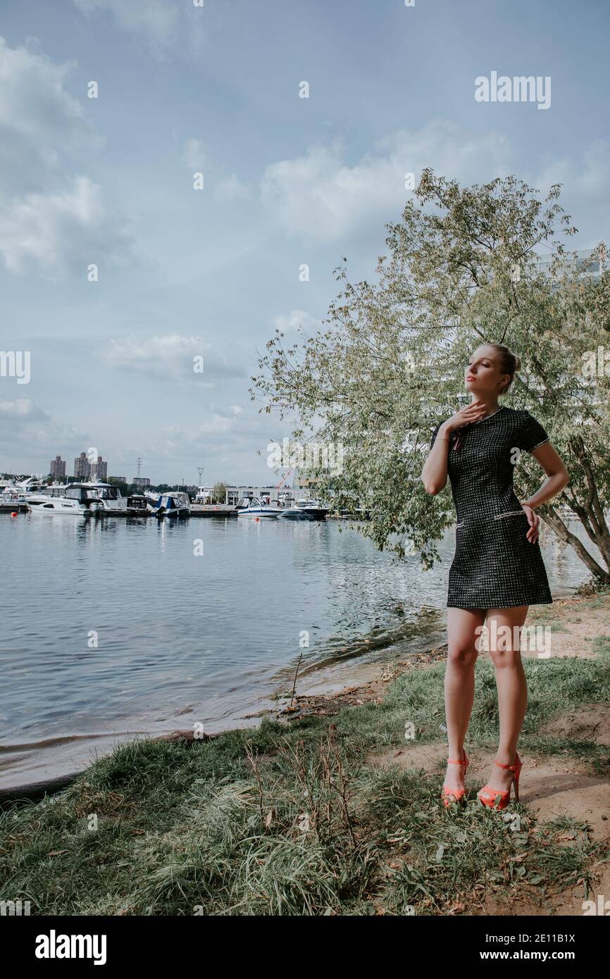 Jolie fille blonde se tient dans le parc près de l'eau avec des bateaux à l'arrière-plan; elle porte une robe courte et élégante sombre et des talons hauts Banque D'Images Jolie fille blonde se tient dans le parc près de l'eau avec des bateaux à l'arrière-plan; elle porte une robe courte et élégante sombre et des talons hauts Banque D'Images