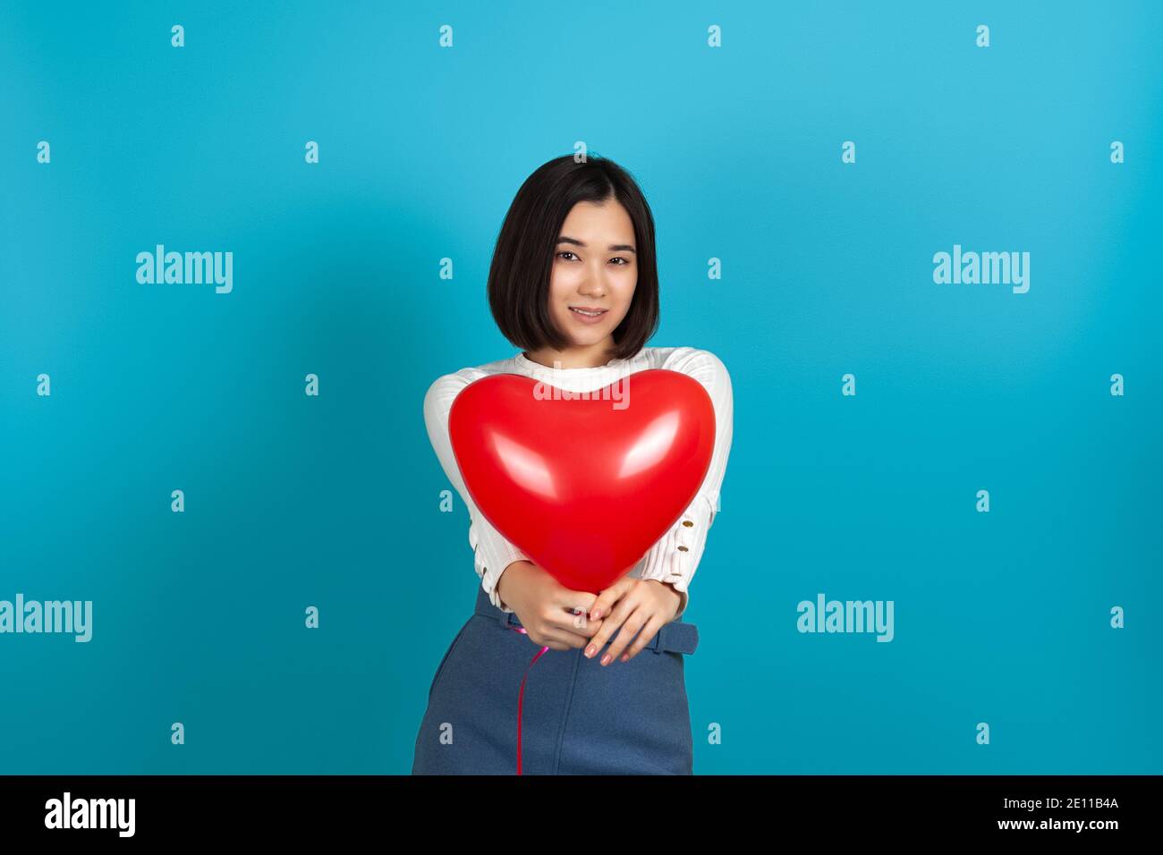 Le modèle d'une femme asiatique souriante présente un rouge ballon en forme de cœur isolé sur fond bleu de la caméra Banque D'Images