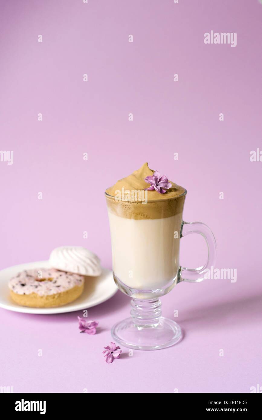 Belle Dalgona buvez un café mousseux dans un mug transparent et des fleurs de lilas sur fond violet. Bonbons sur une assiette: Donat et marshmal tendre Banque D'Images