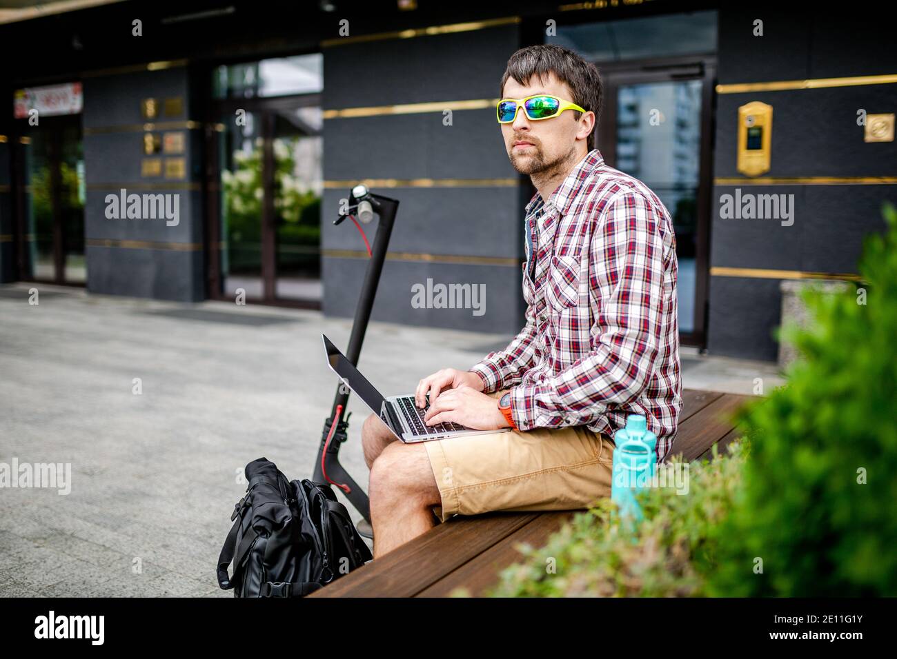 Décontracté et attrayant, le jeune homme de caucasien s'assoit sur un banc à côté de son scooter électrique en utilisant un ordinateur portable pour travailler à distance. Freelance et la technologie moderne font Banque D'Images