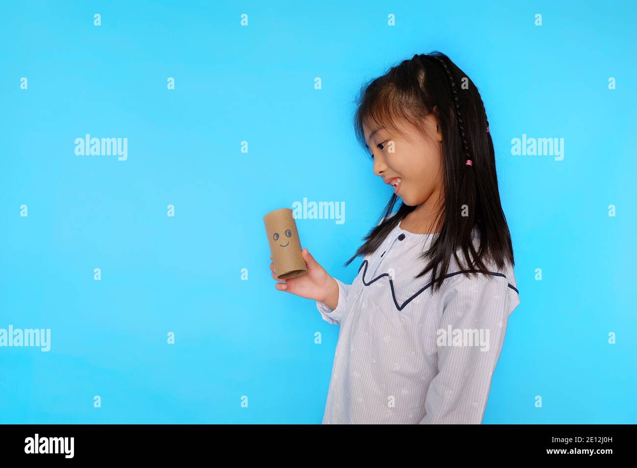 Une jeune fille asiatique mignonne jouant avec un rouleau de tissu vide, dessinant un visage dessus. Fond bleu clair Uni. Banque D'Images