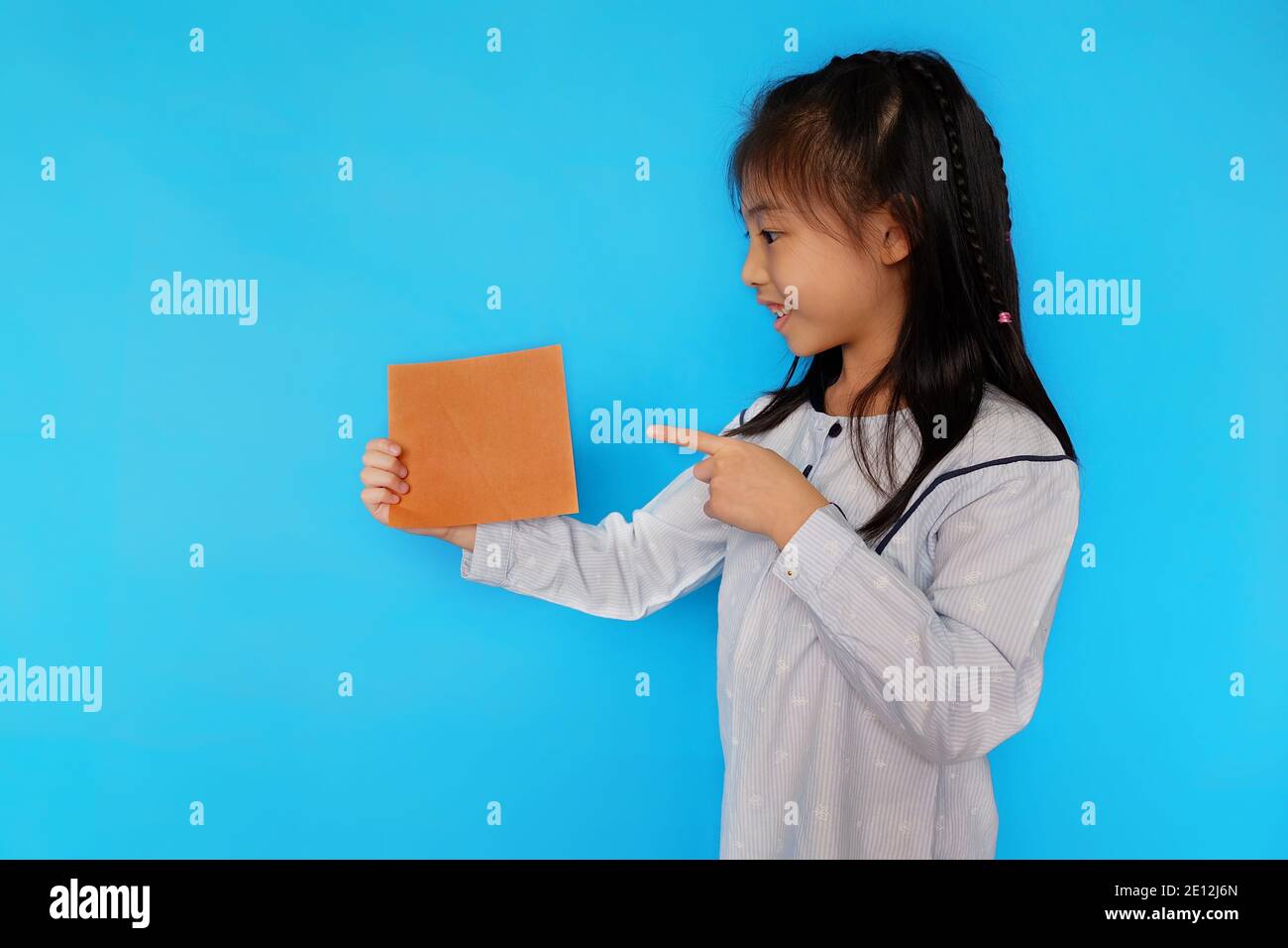 Une jeune fille asiatique mignonne debout sur un fond bleu clair Uni, tenant un morceau de papier vierge, souriant. Banque D'Images