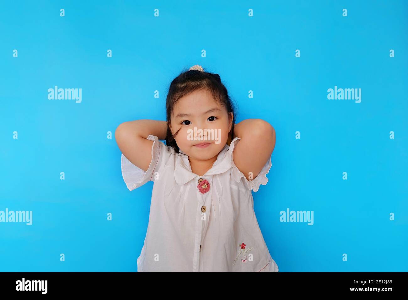 Une jolie jeune fille asiatique confiante est debout avec ses mains derrière sa tête, souriant. Fond bleu clair Uni. Banque D'Images