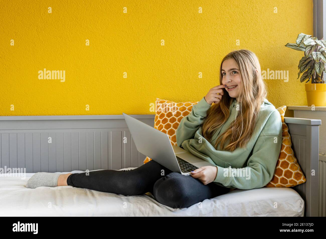Jeune fille blonde utilisant un ordinateur portable sur son lit avant un mur jaune Banque D'Images