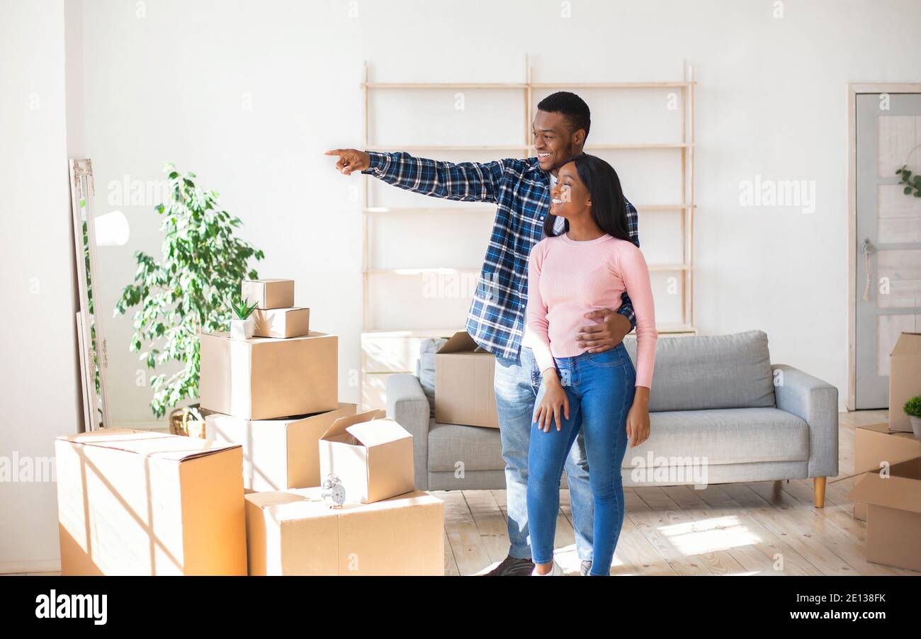 Un gars afro-américain montrant quelque chose à sa copine heureuse, debout parmi les boîtes dans leur nouvelle maison, l'espace libre Banque D'Images