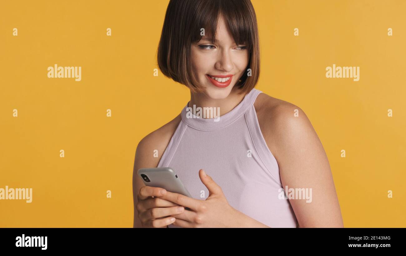 Belle fille de brunette souriante avec bob cheveux rêveur discutant avec petit ami sur smartphone isolé sur fond coloré Banque D'Images