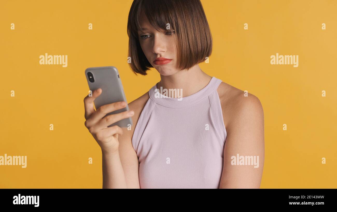 Bouleverser la fille de brunette avec les cheveux de bob regarder triste obtenir le message de l'ami sur smartphone sur fond coloré Banque D'Images