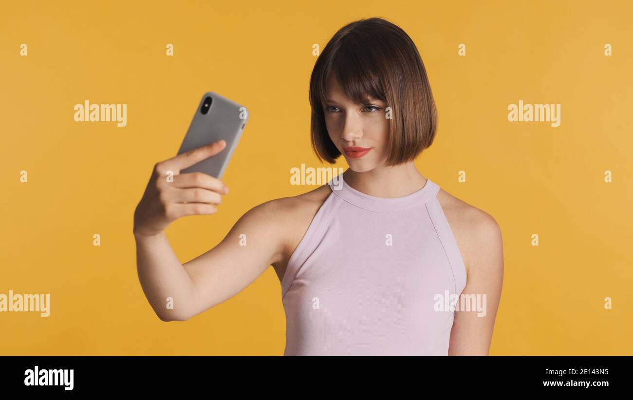 Belle fille brune avec des cheveux de bob et des lèvres rouges faisant selfie sur smartphone sur fond coloré. Concept de technologie moderne Banque D'Images