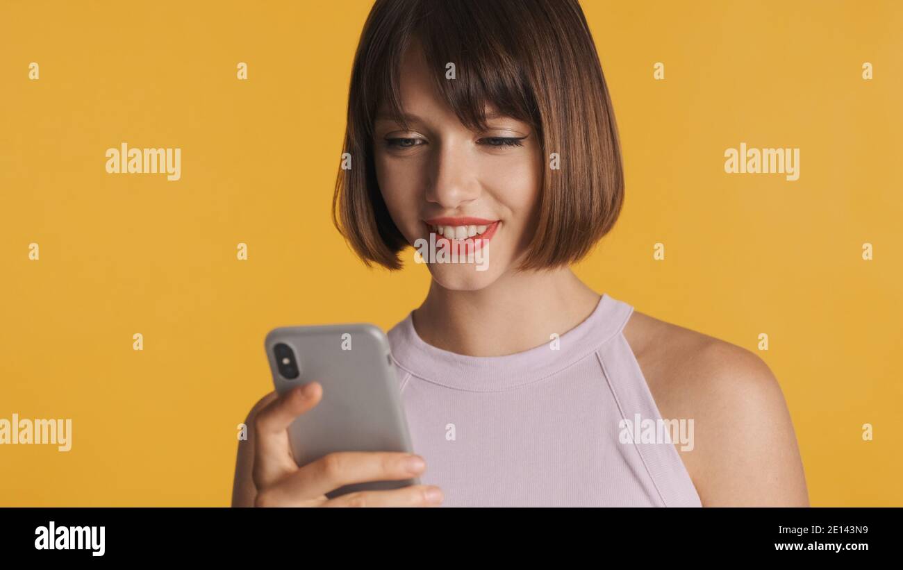Jolie fille brune souriante discutant avec des amis en ligne sur smartphone isolé sur fond coloré Banque D'Images