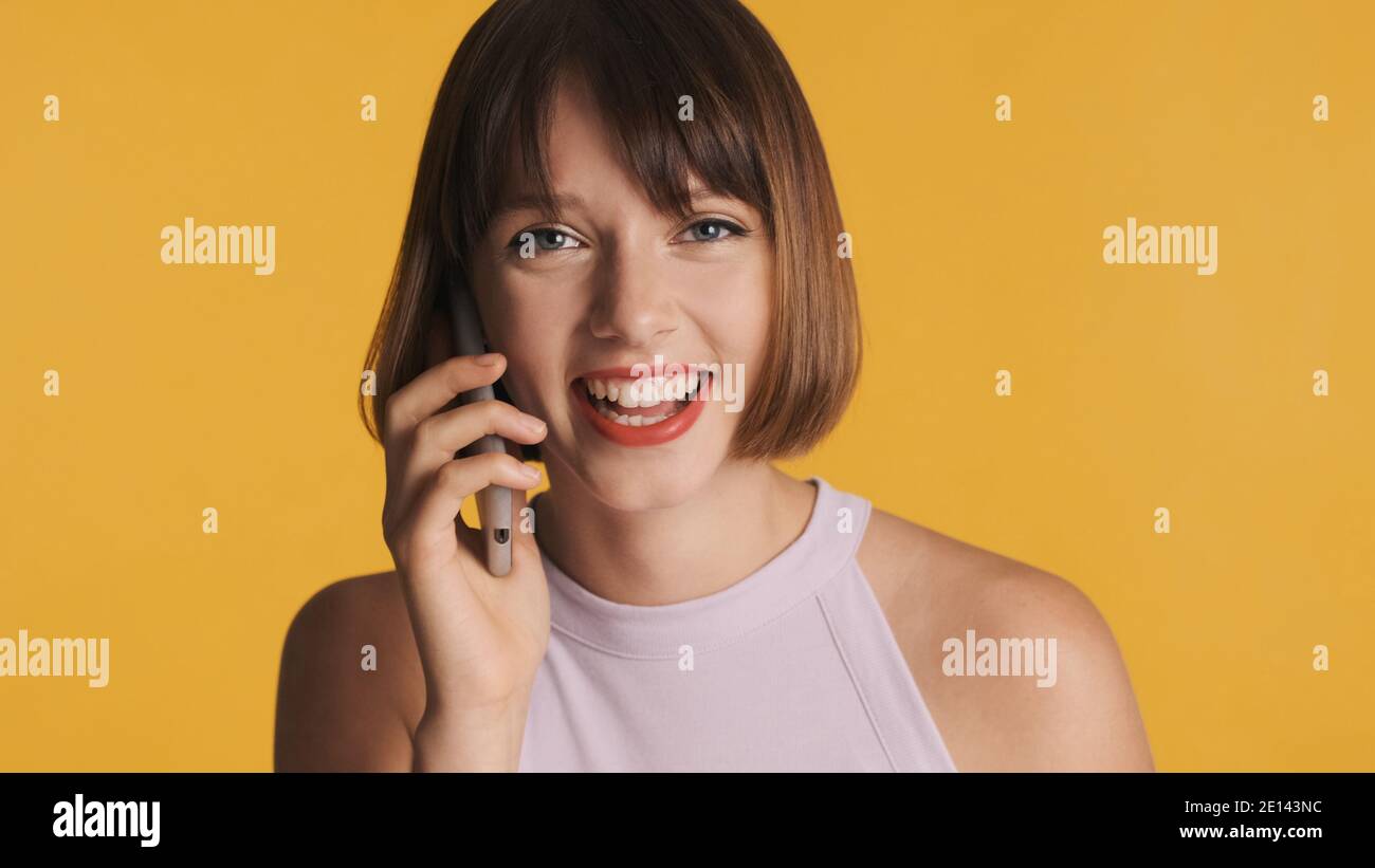 Belle fille gaie de brunette avec bob cheveux se réjouissant de parler avec ami sur smartphone isolé sur fond jaune Banque D'Images