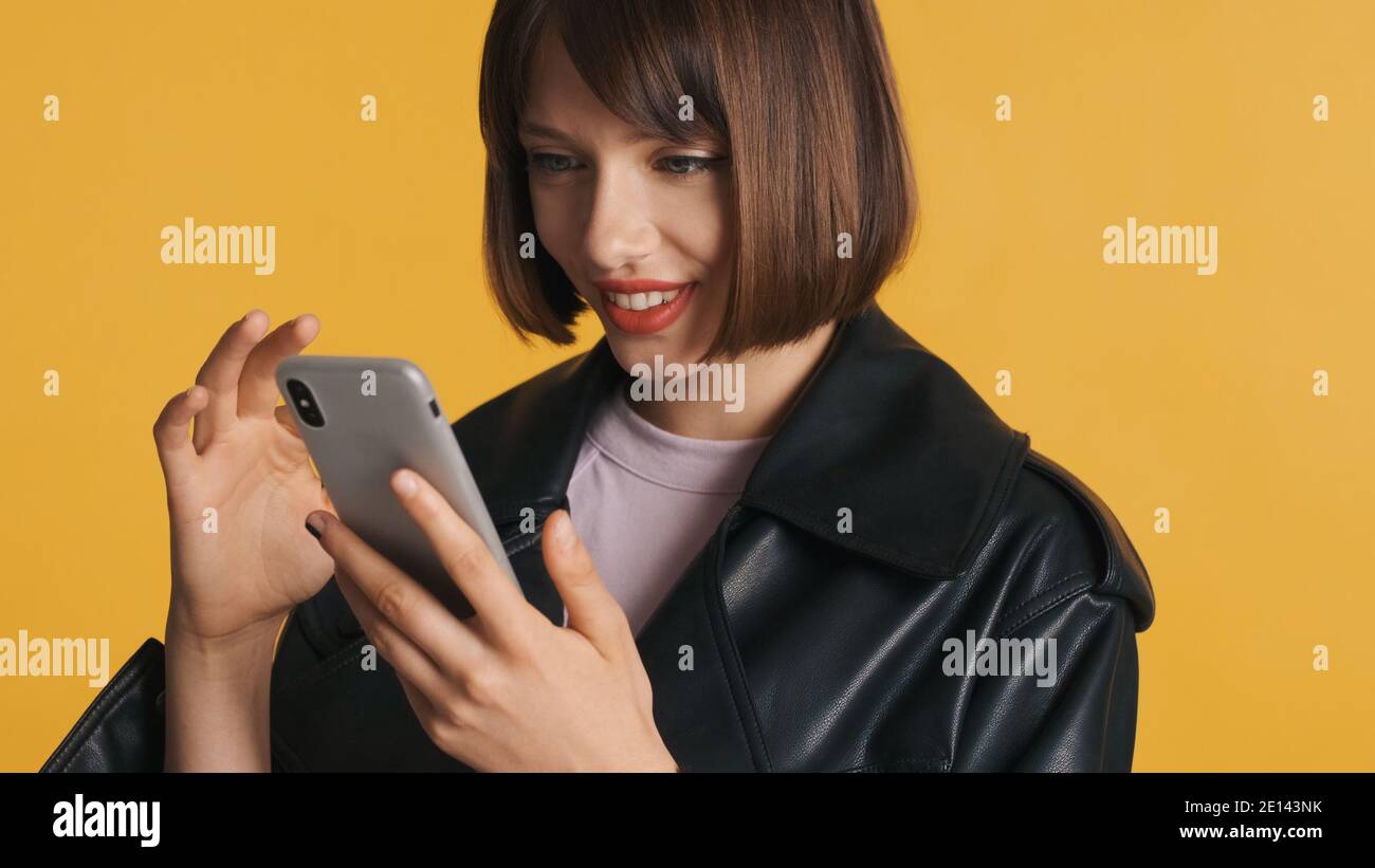 Portrait d'une jolie fille brunette avec des cheveux de bob dans une ambiance décontractée portez joyeusement les médias sociaux surfuges sur un smartphone isolé sur jaune arrière-plan Banque D'Images