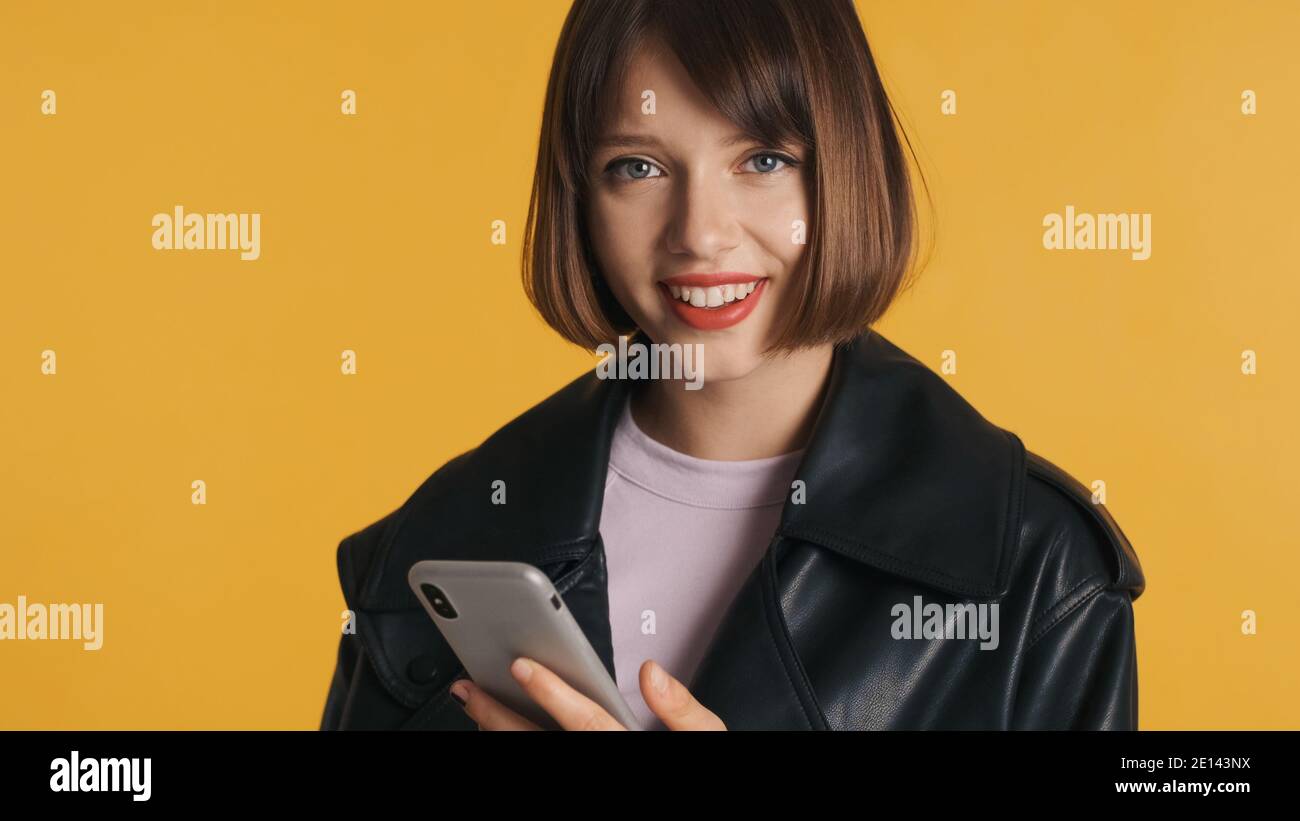 Belle fille de brunette avec bob cheveux en utilisant le smartphone heureusement regarder dans l'appareil photo isolé sur fond jaune Banque D'Images