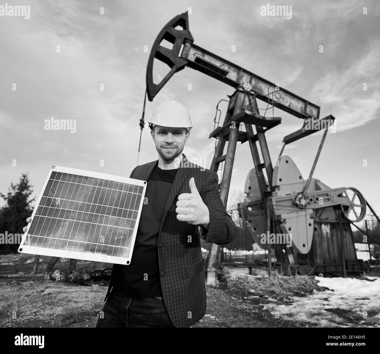 Homme d'affaires tenant un mini-module solaire et montrant le pouce vers le haut, debout dans le champ de pétrole à côté de l'engin de forage pétrolier. Concept de l'industrie pétrolière, sources d'énergie alternatives. Image en noir et blanc Banque D'Images