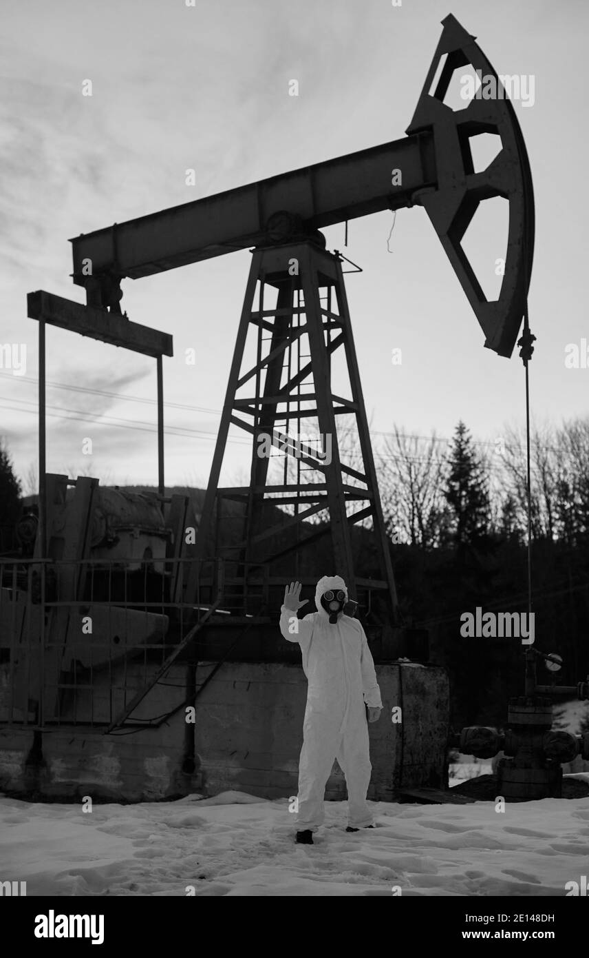 Scientifique dans le masque à gaz debout sur le territoire du champ de pétrole avec la machine à bascule de pompe à huile sur le fond. Le chercheur montre le panneau stop devant le cric de la pompe à huile. Concept d'écologie. Image en noir et blanc Banque D'Images