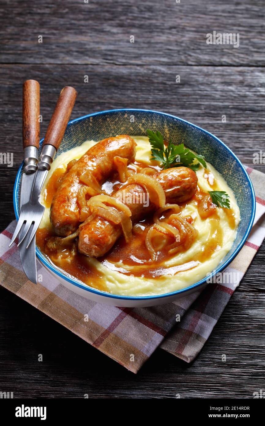 Bangers et purée de saucisses britanniques traditionnelles de porc avec sauce à l'oignon et purée de pommes de terre sur une assiette bleue avec des couverts sur un fond en bois sombre, Banque D'Images