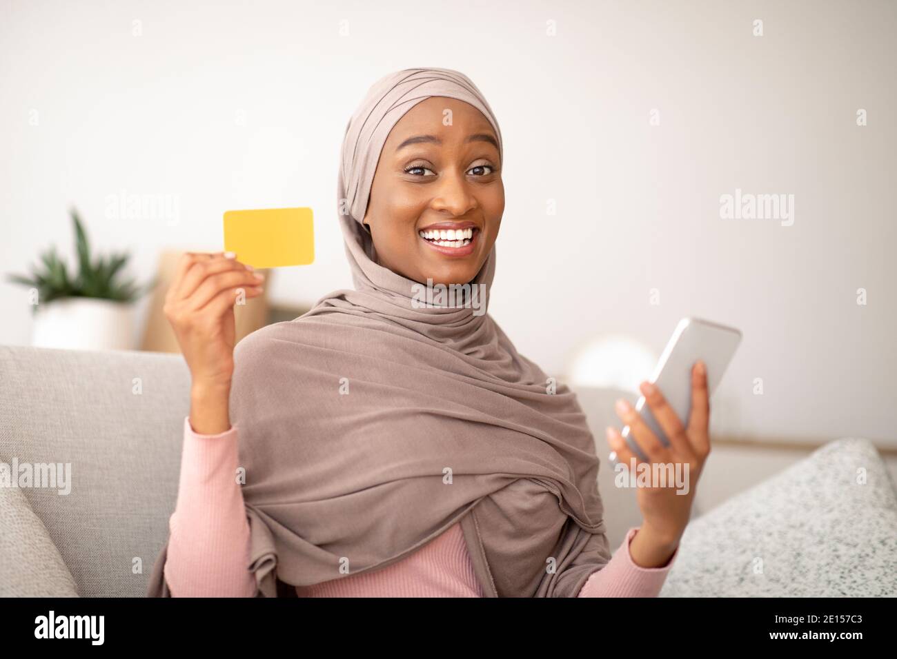 Bonne femme noire dans hijab tenant la carte de crédit et le téléphone cellulaire, achetant des marchandises en ligne par application à la maison Banque D'Images