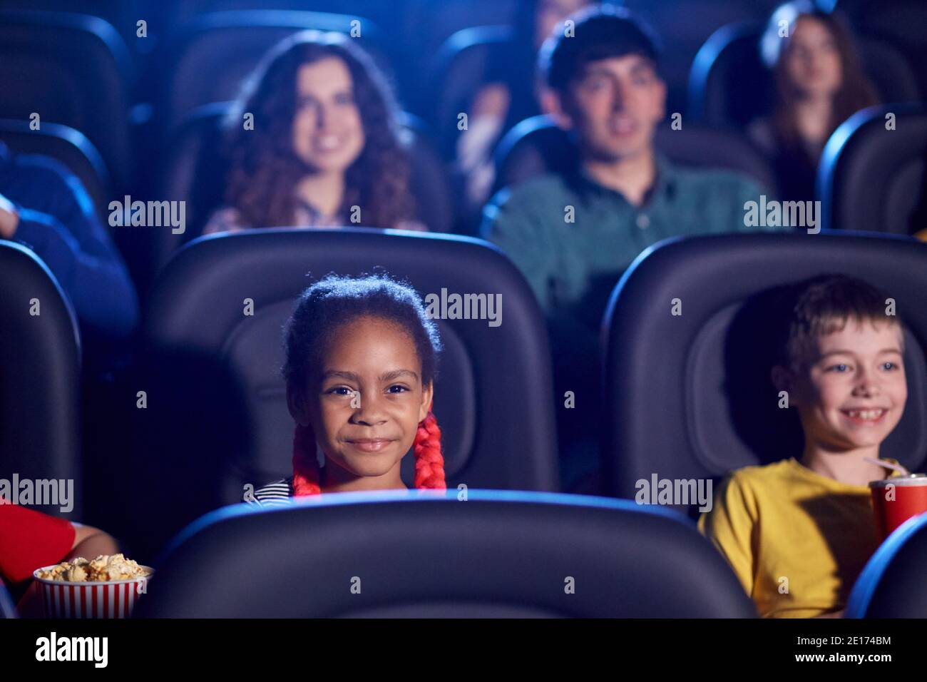 Vue de face des enfants multiraciaux en train de passer du temps ensemble, un public jeune en arrière-plan. Foyer sélectif de la petite fille africaine heureuse assise dans le cinéma, regardant le dessin animé drôle. Concept de divertissement. Banque D'Images