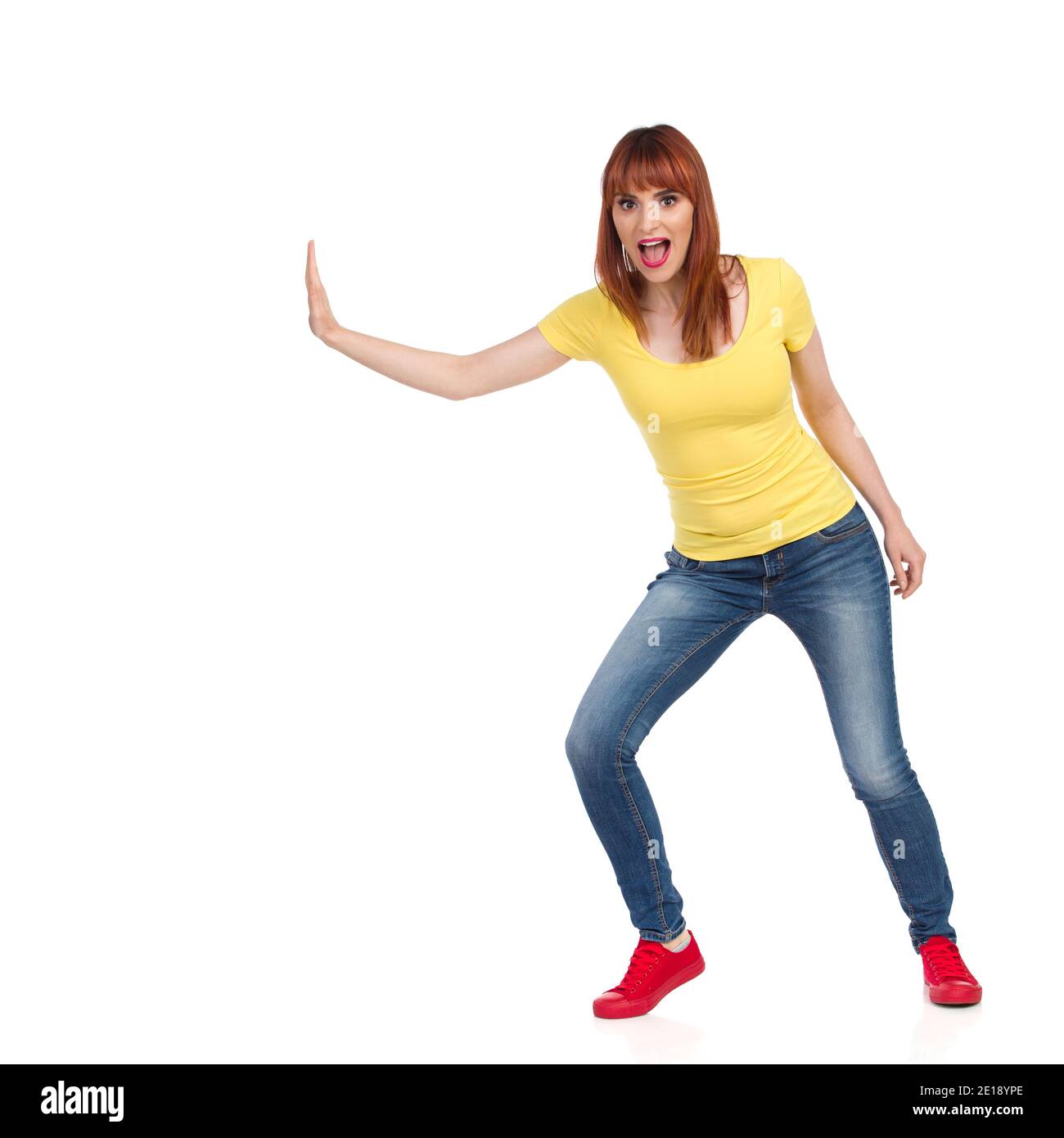 Une jeune femme excitée en chemise jaune, jeans et baskets rouges pousse quelque chose d'une main et crie. Vue avant. Prise de vue en studio pleine longueur isola Banque D'Images