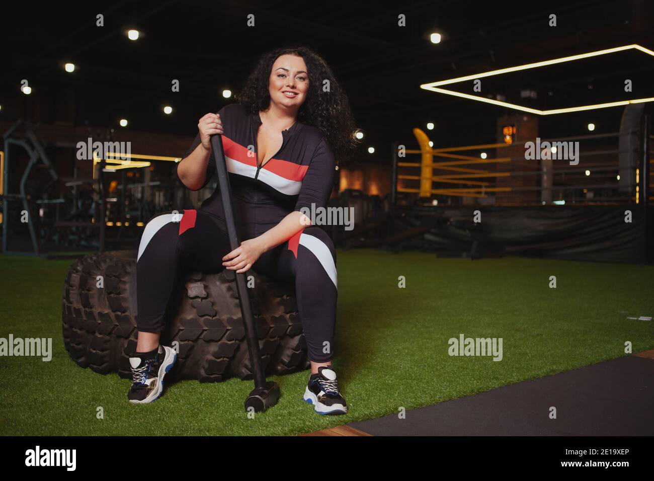 Femme pleine de confiance et de taille souriant à l'appareil photo, assise sur un pneu de roue lourd à la salle de gym, tenant un marteau de traîneau, un espace de copie. Joyeux sport en surpoids Banque D'Images